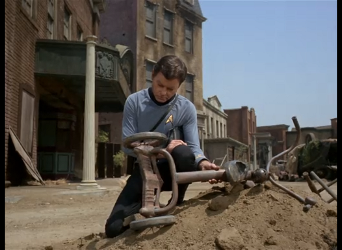 McCoy kneeling in a dusty street in front of some dilapidated buildings, looking at an old, broken tricycle. 
