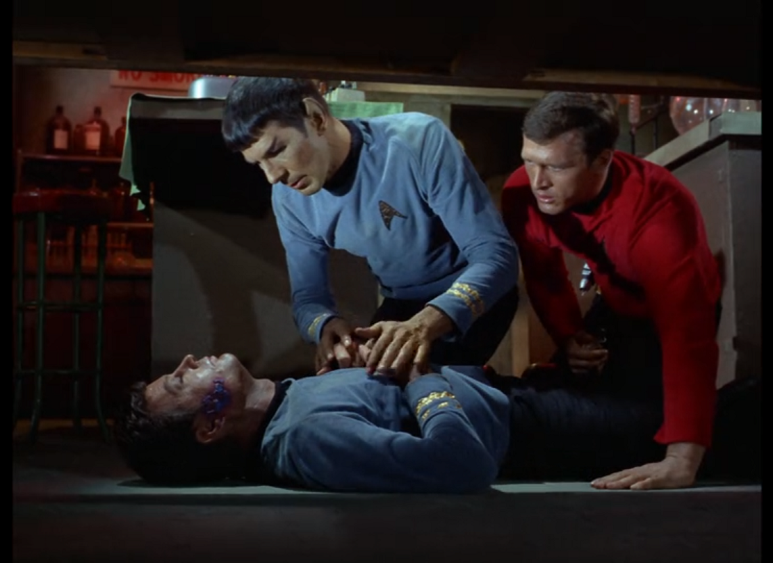 McCoy laying unconscious on the floor, while Spock and a redshirt kneel over him, Spock gripping McCoy's hands in his. 